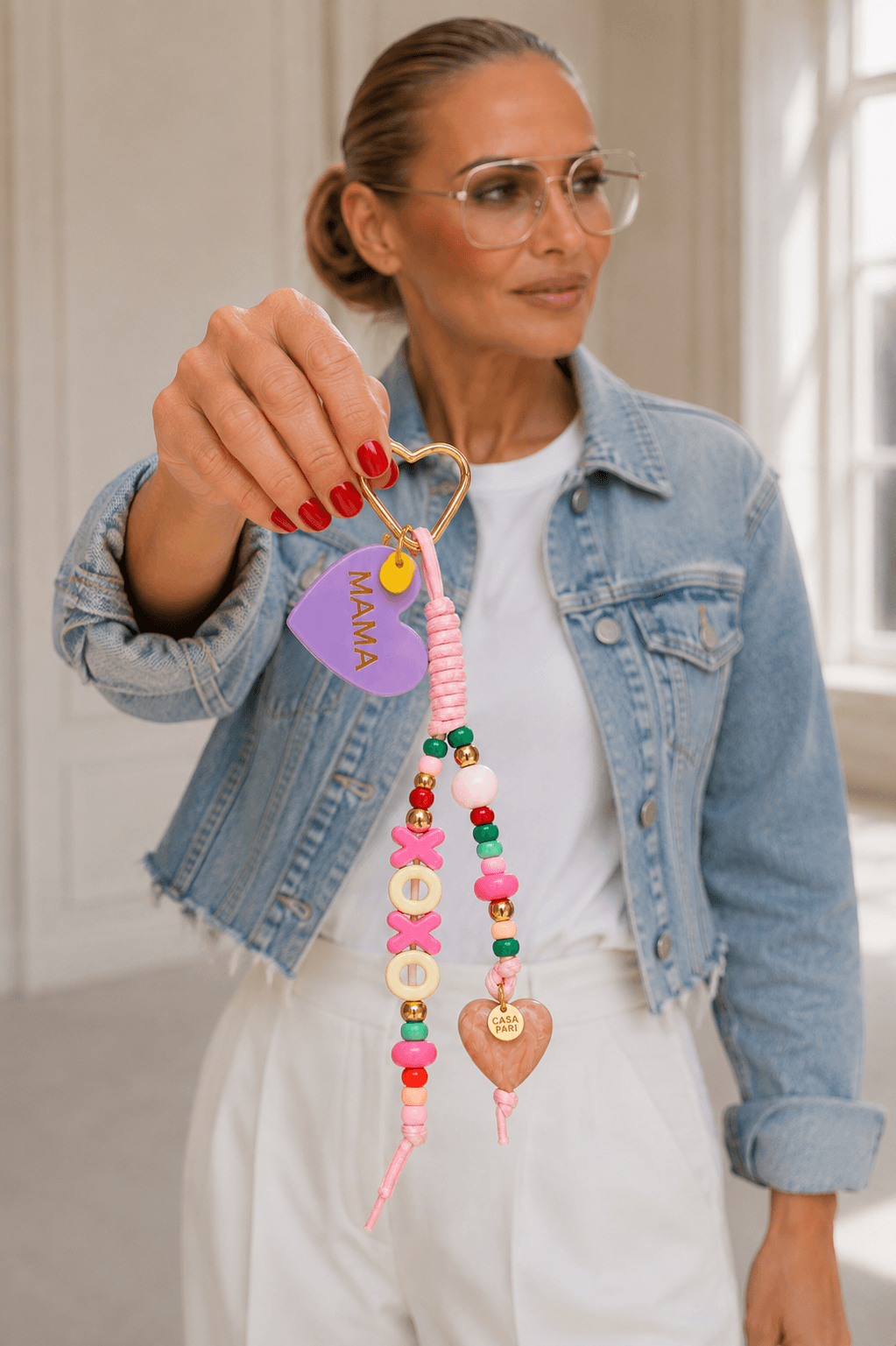 MAMA Bag Charm von CASAPARI mit lilafarbenem Herz, XOXO Buchstaben, bunten Perlen und goldfarbenem Herz-Karabiner, von einem Model in Jeansjacke präsentiert.