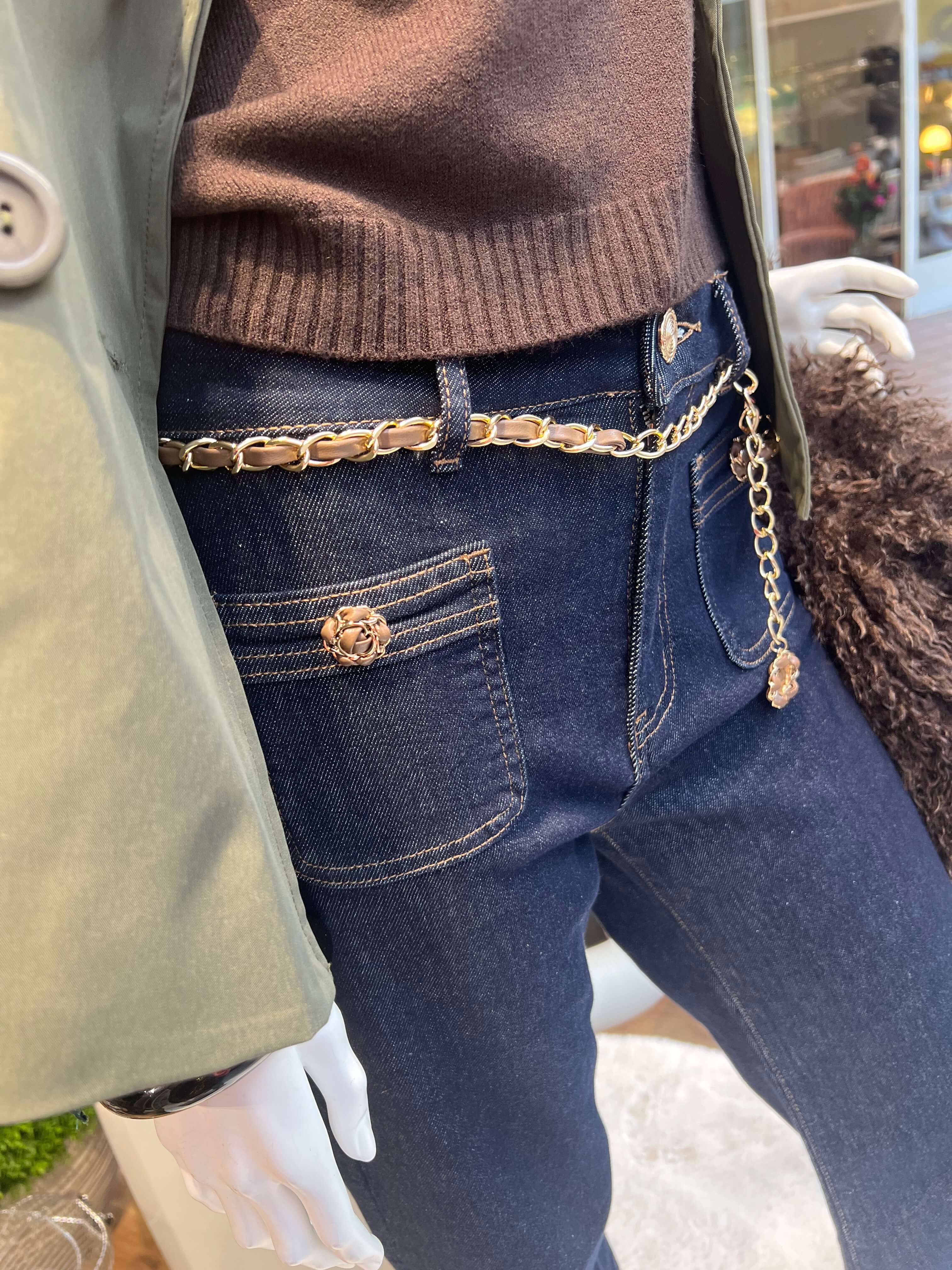 Detail der dunkelblauen Marlene Jeans mit goldfarbenem Kettengürtel, eingeflochtenem Leder-Look und Blütenknopf.