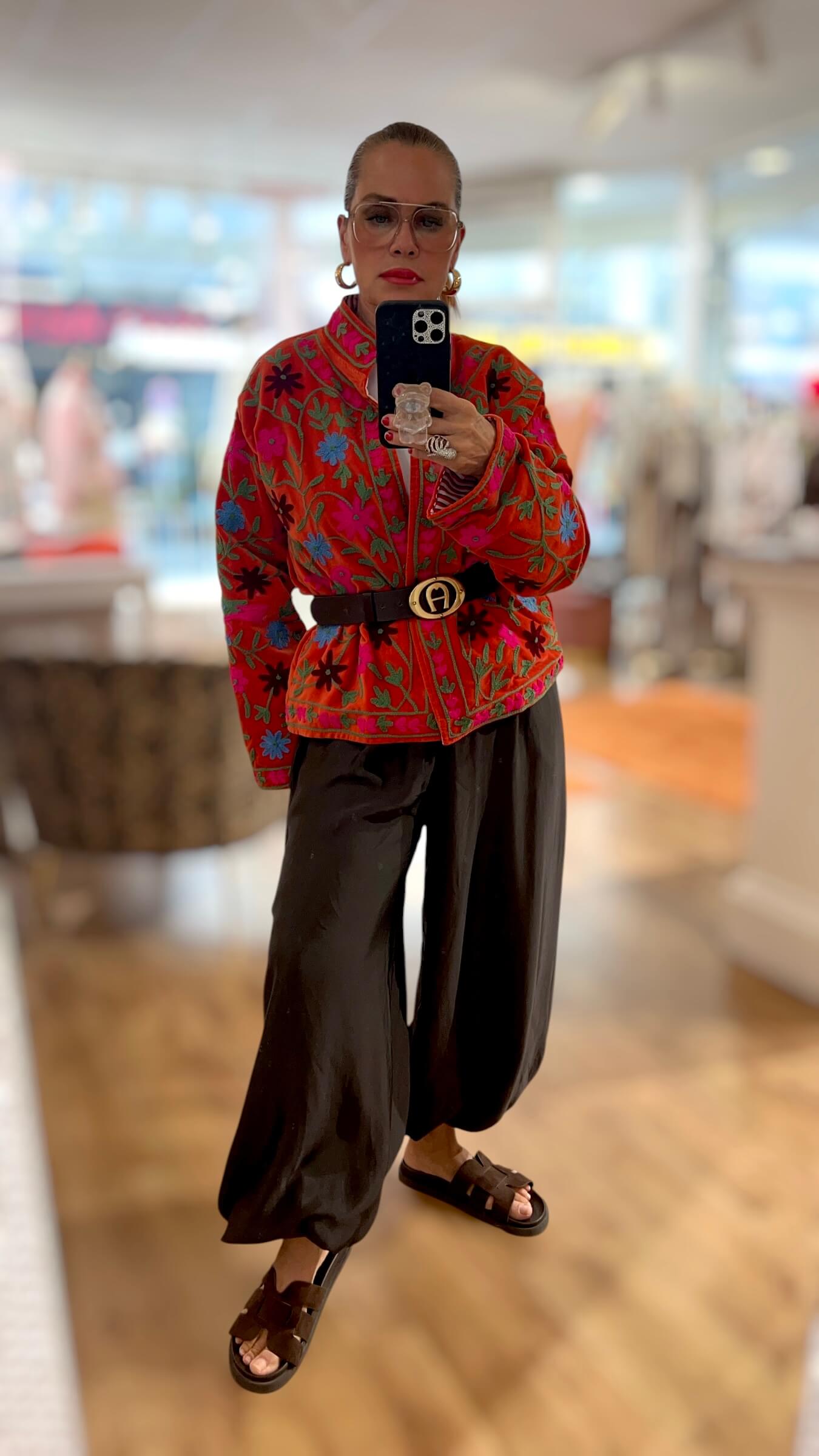 Rote Couture-Jacke mit aufwendiger floraler Stickerei in Pink, Blau und Grün. Die Jacke wird mit Gürtel zu einer weiten schwarzen Hose kombiniert.