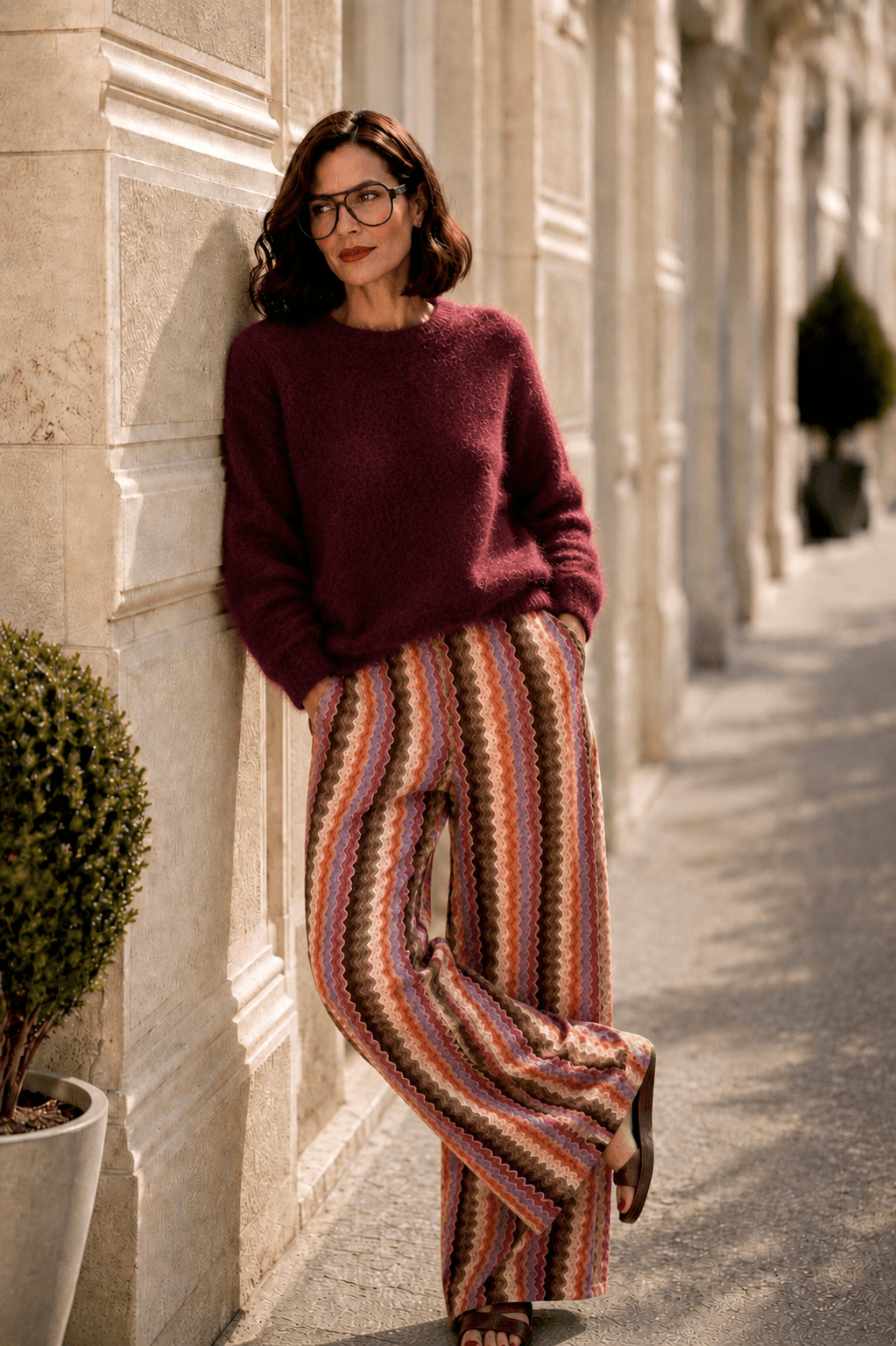 Frau trägt einen flauschigen Milano Kidmohair Pullover in Burgundy kombiniert mit einer gestreiften Marlenehose und lehnt an einer hellen Natursteinfassade.