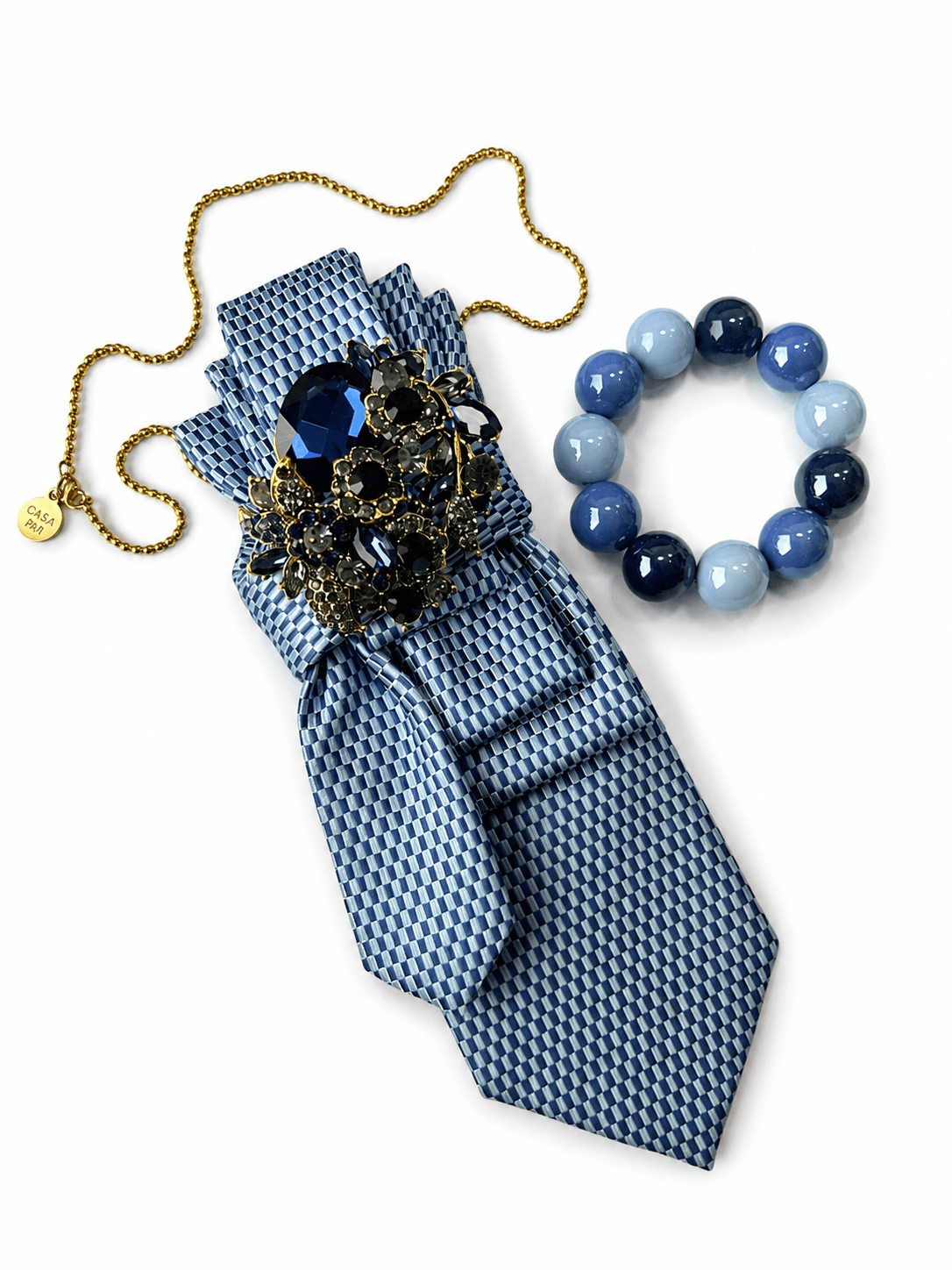 CASAPARI Perfect Match Affair Blue Hour mit blau gemusterter Krawatte, Schmuckbrosche und farblich abgestimmtem Perlenarmband in verschiedenen Blautönen.