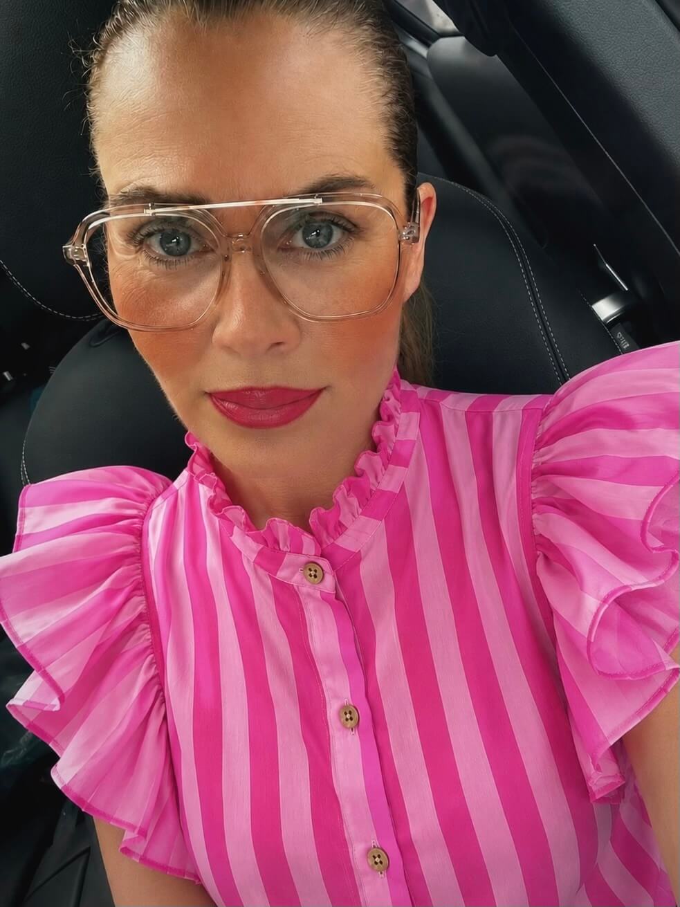 Frau in Nahaufnahme im Auto trägt eine ärmellose rosa-gestreifte Bluse mit Stehkragen und großen Rüschen an den Schultern, transparente Brille, pinken Lippenstift und blaue sowie pinke Ohrringe.