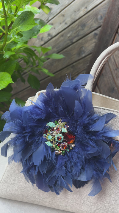 Blaue Brosche mit Schmuckelement an einer Beigen Tasche drapiert. Hinter einer Holzwand