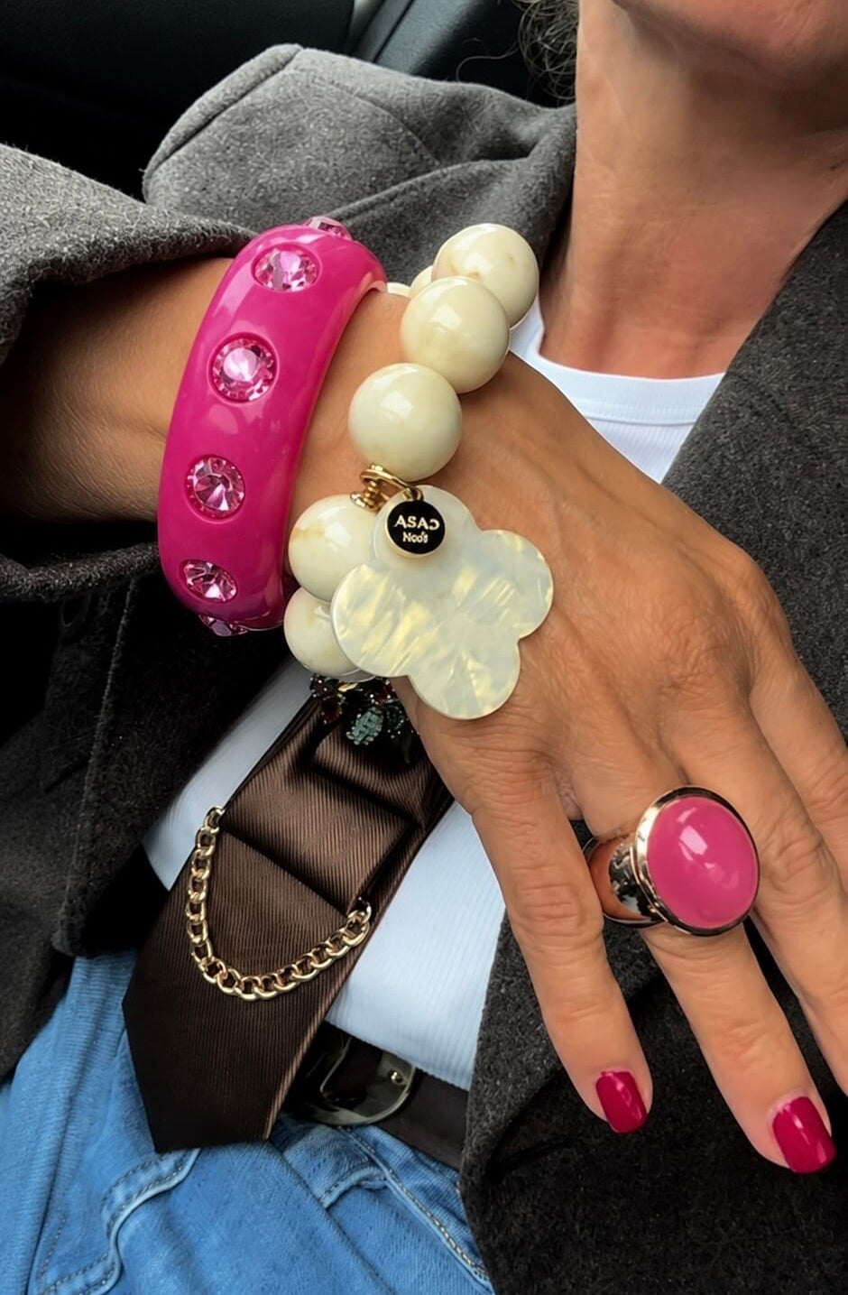 Handgelenk mit pinkfarbenem Resina-Armreif mit Strass-Steinen, kombiniert mit einem cremefarbenen Perlenarmband und einem pinken Ring. Die Person trägt einen grauen Blazer, Jeans und eine braune Krawatte mit goldfarbener Kette.