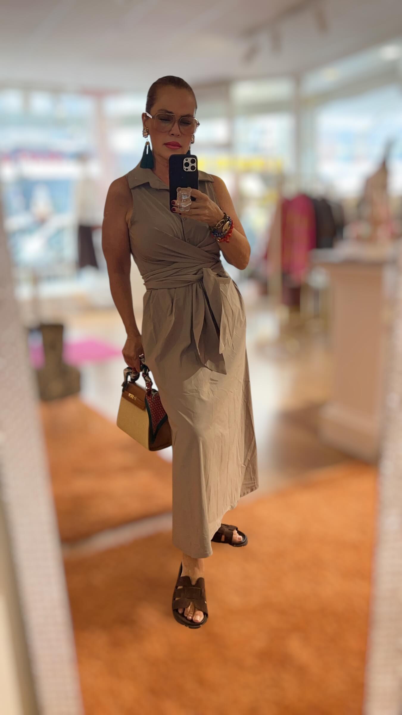 Frau trägt ein ärmelloses Wickelkleid in Greige mit Hemdkragen und Bindegürtel. Das knöchellange Kleid hat eine fließende Silhouette und wird mit dunklen Sandalen, Statement-Ohrringen und einer kleinen Handtasche kombiniert. Fotografiert in einer hellen Boutique.